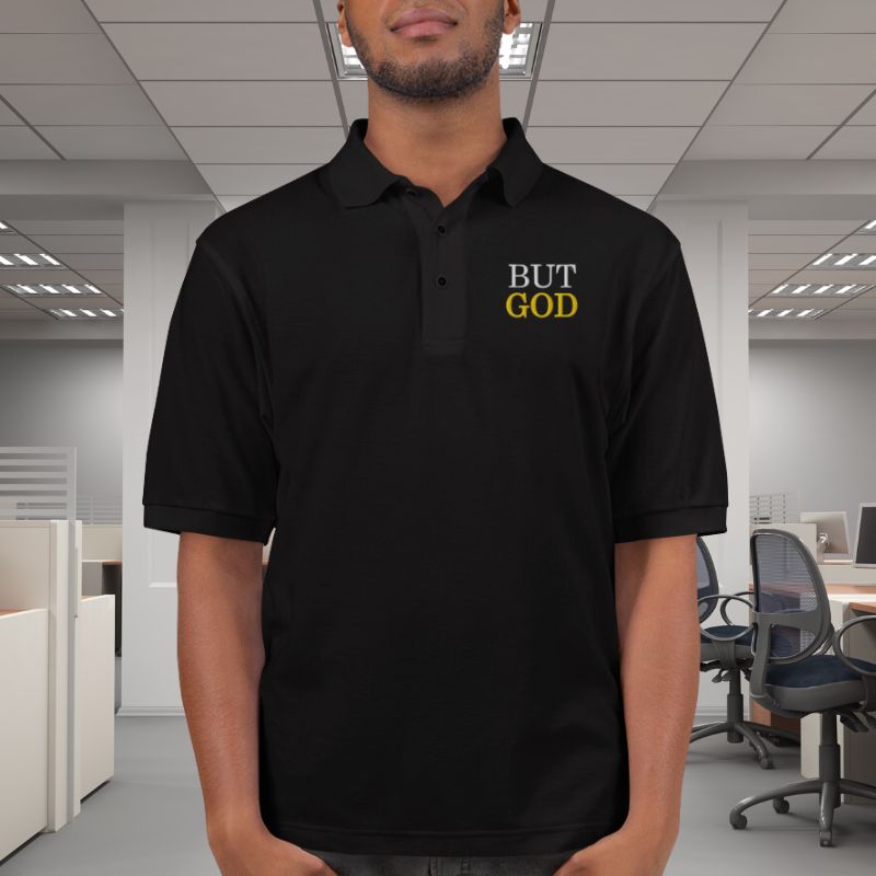 Hispanic man wearing a black polo shirt. On the top left corner, there is an embroidered design that says &quot;BUT GOD&quot; in caps lock. This shirt is sold at Amela&