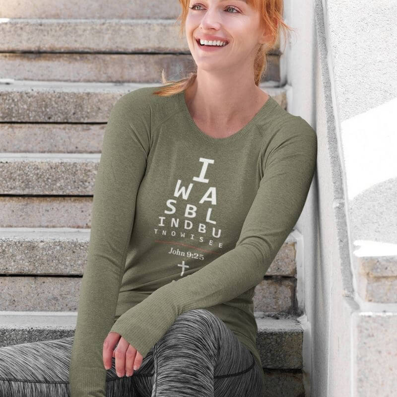 White woman wearing a long-sleeve sage green Christian shirt. 

The graphic is text as an eye chart, and it says &quot;I WAS BLIND BUT NOW I SEE&quot;.

Below the text, there is a thin line, with text &quot;John 9:25&quot; and an icon of a cross below.