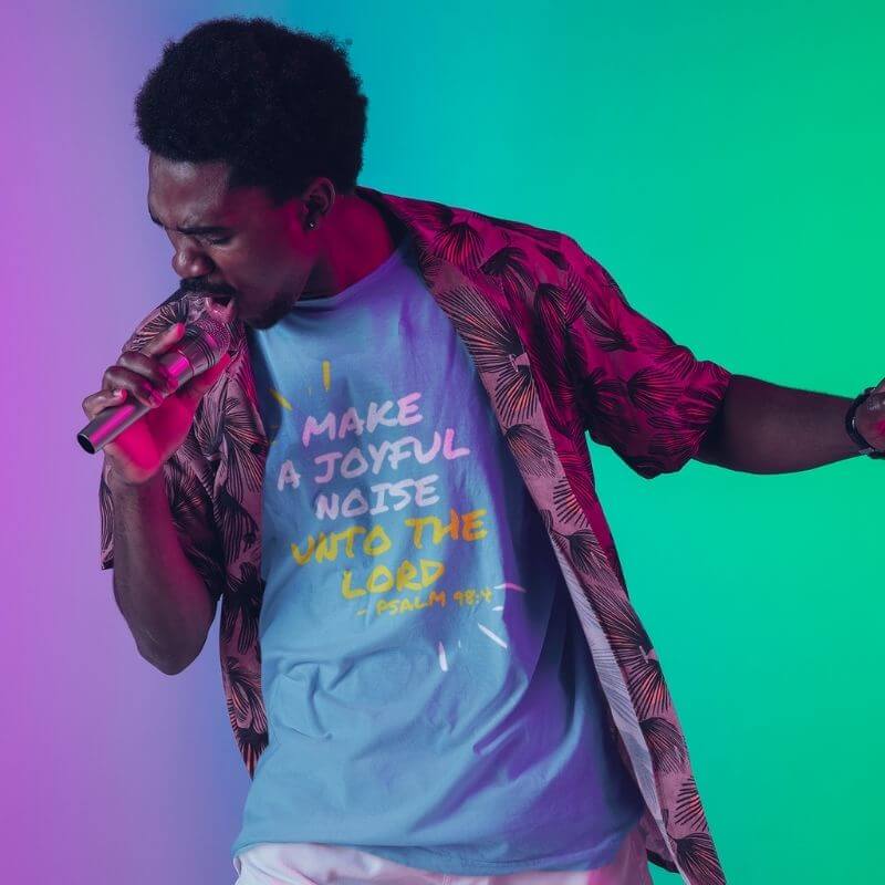 Black man singing into a microphone while wearing and showcasing a blue Christian t-shirt that says &quot;Make joyful noise unto the Lord&quot; with &quot;Pslam 98:4 underneath&quot;. Surrounding the text, there are lines that show movement or music.