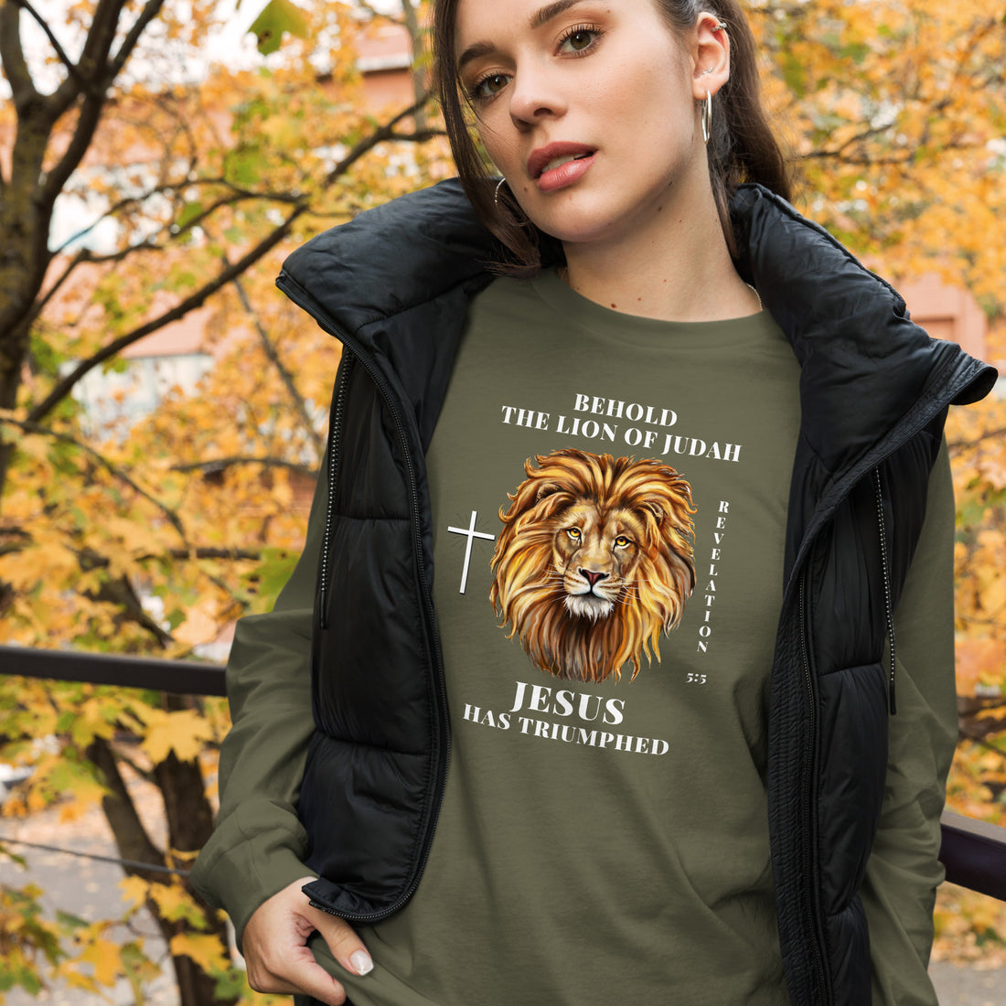 White woman wearing an olive green shirt that says &quot;Behold the lion of Judah&quot;. Below the text, there is a graphic of a lion, with a cross icon next to it, and &quot;Revelation 5:5&quot;. On the bottom, text says &quot;JESUS has triumphed&quot;