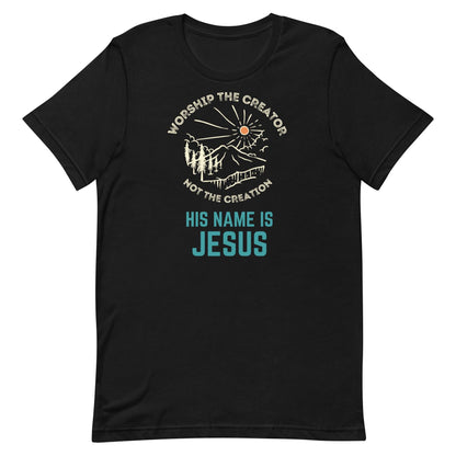 Flatlay of a black Christian t-shirt. There is a graphic of a mountain, trees, and sun. Over the graphic, it says &quot;Worship the creator&quot;, and under it says &quot;not the creation&quot;. On the bottom, it says &quot;His name is Jesus&quot;. This shirt is sold at Amela&