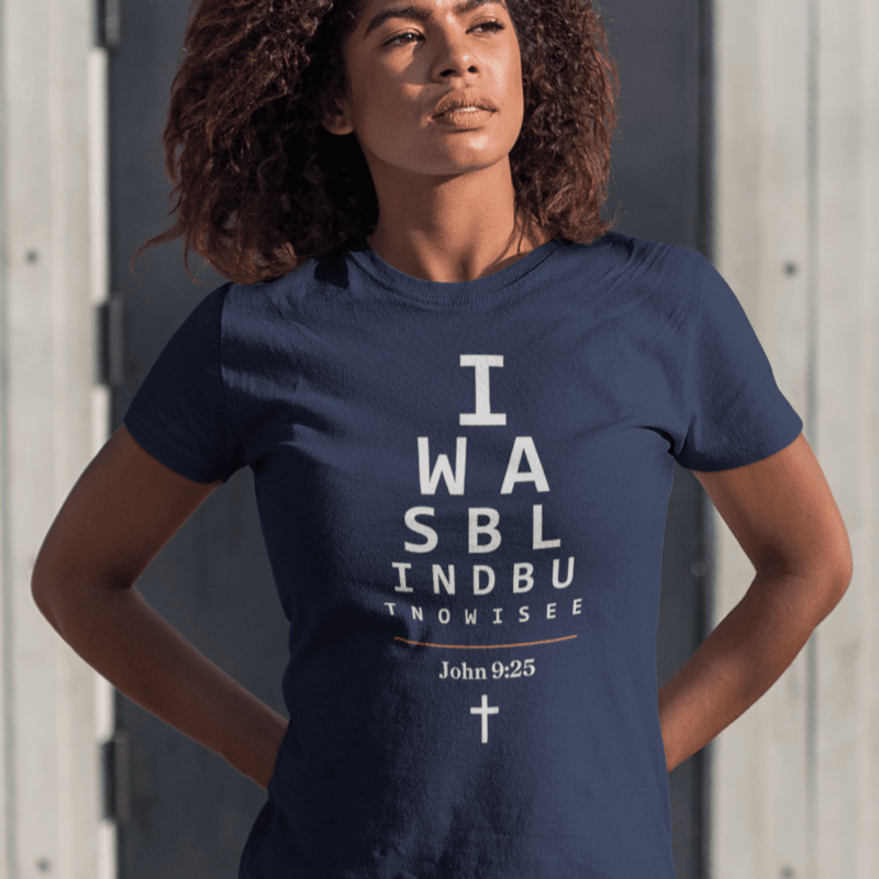 Black woman wearing a navy Christian t-shirt. 

The graphic is text as an eye chart, and it says &quot;I WAS BLIND BUT NOW I SEE&quot;.

Below the text, there is a thin line, with text &quot;John 9:25&quot; and an icon of a cross below.