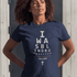 Black woman wearing a navy Christian t-shirt. 

The graphic is text as an eye chart, and it says "I WAS BLIND BUT NOW I SEE".

Below the text, there is a thin line, with text "John 9:25" and an icon of a cross below.